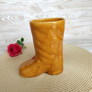 May include: A ceramic boot-shaped vase in a brown glaze. The vase is sitting on a woven placemat.