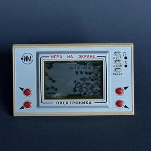 Könnte beinhalten: Eine beige tragbare elektronische Spielkonsole mit einem rechteckigen Bildschirm, der eine Digitaluhr und Spielgrafiken anzeigt. Das Gerät verfügt über vier rote runde Tasten, eine silberne Frontplatte mit kyrillischem Text und ein IM-Logo.