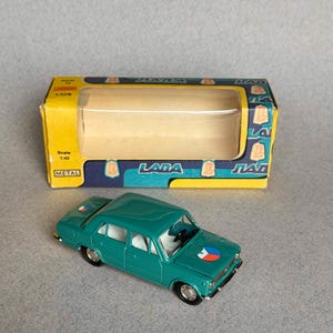 Puede incluir: Un coche modelo LADA de color azul turquesa en una caja. El coche tiene un interior blanco y una pegatina de la bandera checa en el capó. La caja es azul y amarilla con la palabra "LADA" impresa en ella.