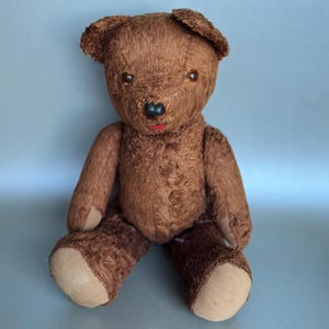 May include: A vintage brown teddy bear with a red mouth and tan paws. The bear has button eyes and a classic design. The plush toy is sitting upright, with its arms and legs extended.
