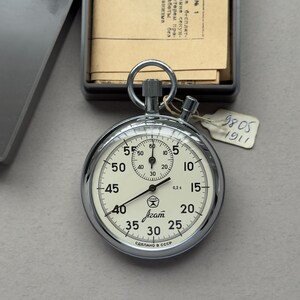 May include: A chrome-plated mechanical stopwatch with a cream-colored dial, black numerals, and a small sub-dial. The face features the text Agat, a star logo, and the Cyrillic text СДЕЛАНО В СССР. A small white tag with the handwritten numbers 9805 1911 is attached.