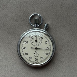 Può includere: Un cronometro vintage con cassa argentata e quadrante bianco. Il quadrante presenta numeri e segni neri per secondi e minuti. Il cronometro ha un piccolo quadrante in alto e un pulsante laterale. Il testo "MADE IN USSR" è visibile.