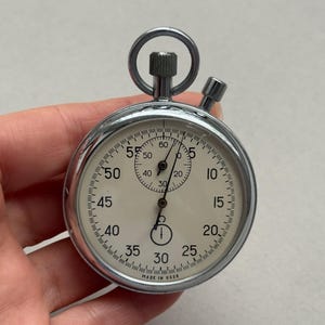 May include: A vintage silver-tone mechanical stopwatch with a cream-colored face, black numerals, and two sub-dials. The device features a top winding crown with a circular ring and a side pusher. The text "MADE IN USSR" is printed at the bottom.