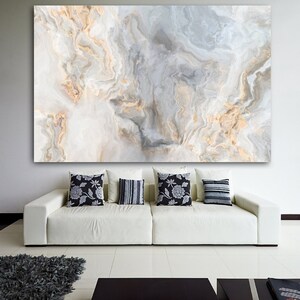 May include: A white couch with black and white patterned throw pillows in front of a large abstract painting with gray and gold swirls. The painting is on a white wall in a modern living room.