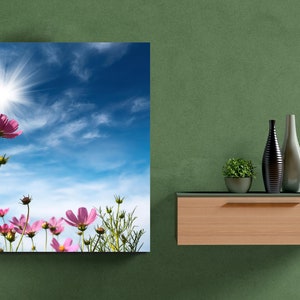 K&ouml;nnte beinhalten: Quadratischer Leinwanddruck mit rosa Kosmosbl&uuml;ten vor strahlender Sonne und blauem Himmel. Das Kunstwerk h&auml;ngt an einer gr&uuml;nen Wand, mit dekorativen Vasen und einer kleinen Pflanze auf einem Holzregal.