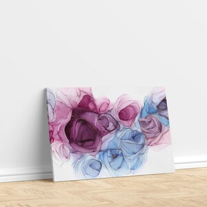 May include: Canvas art print featuring an abstract floral design. The artwork showcases a cluster of stylized roses in shades of pink, purple, and blue, with gold accents. The canvas is leaning against a white wall on a wooden floor.