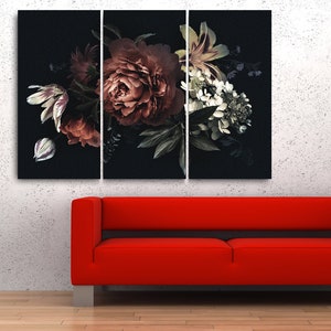 Op de afbeelding: Een drieluik canvas print met een donker bloemstuk met bordeauxrode, witte en groene bloemen tegen een zwarte achtergrond. Een rode bank en zwarte vaas met takken staan op de voorgrond.
