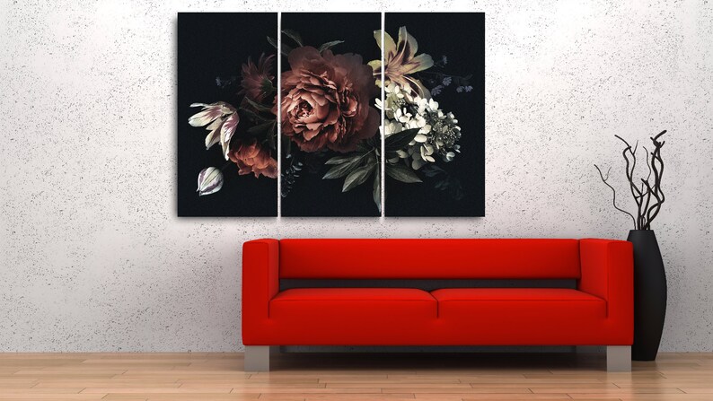 Op de afbeelding: Een drieluik canvas print met een donker bloemstuk met bordeauxrode, witte en groene bloemen tegen een zwarte achtergrond. Een rode bank en zwarte vaas met takken staan op de voorgrond.