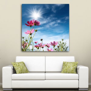 K&ouml;nnte beinhalten: Ein Leinwanddruck mit rosa Kosmosbl&uuml;ten vor einem strahlend blauen Himmel mit einer strahlenden Sonne. Das Kunstwerk h&auml;ngt &uuml;ber einem wei&szlig;en Sofa mit gr&uuml;n gemusterten Kissen, flankiert von Topfpflanzen.