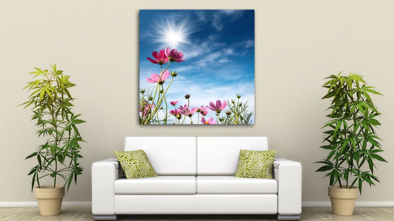 K&ouml;nnte beinhalten: Ein Leinwanddruck mit rosa Kosmosbl&uuml;ten vor einem strahlend blauen Himmel mit einer strahlenden Sonne. Das Kunstwerk h&auml;ngt &uuml;ber einem wei&szlig;en Sofa mit gr&uuml;n gemusterten Kissen, flankiert von Topfpflanzen.