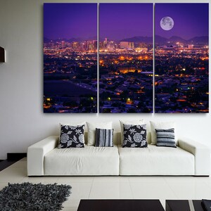 May include: A panoramic view of a city skyline at night with a large, bright moon in the sky. The city is lit up with lights, and the buildings are silhouetted against the purple sky.