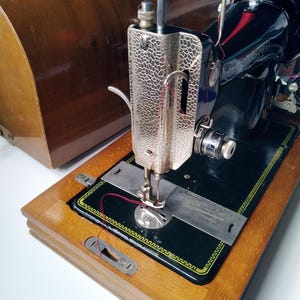 May include: Vintage black sewing machine with a silver-toned faceplate and a wooden base. A spool of red thread is visible. The machine is in a wooden case. The sewing machine is on a white surface.