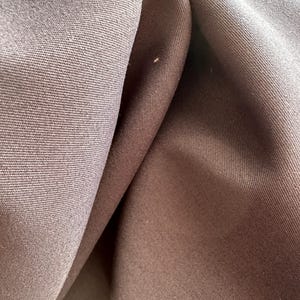May include: A close-up view of dark brown fabric featuring a tight, uniform weave. The material is draped to create soft, deep folds and shadows, highlighting the consistent texture and matte finish of the textile surface across the entire frame.