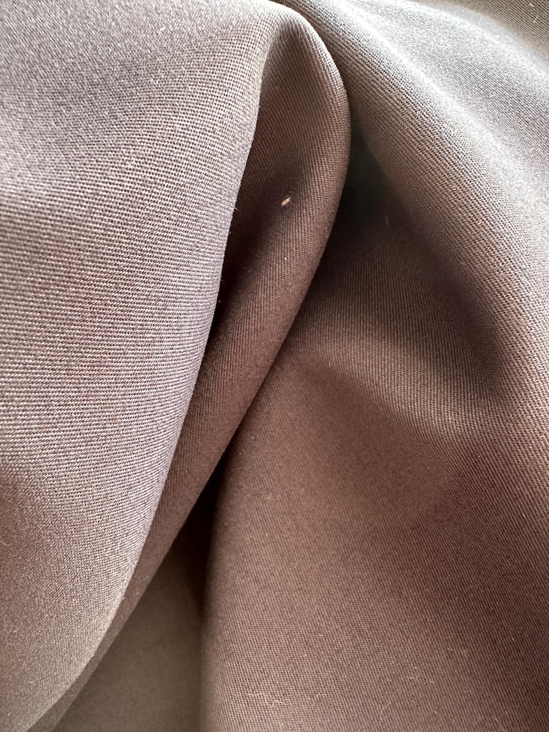 May include: A close-up view of dark brown fabric featuring a tight, uniform weave. The material is draped to create soft, deep folds and shadows, highlighting the consistent texture and matte finish of the textile surface across the entire frame.