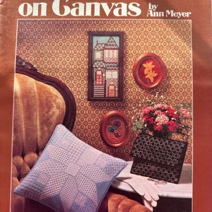 May include: A vintage craft book cover featuring a pulled thread on canvas project. The cover shows a blue and white pillow with a star design, a framed picture of a house, and a small framed picture of a leaf. The title of the book is "Pulled Thread on Canvas" by Ann Meyer. The book is from Leisure Arts and is leaflet 75.