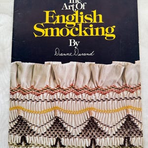 May include: A vintage book titled "The Art of English Smocking" by Dianne Durand. The cover is black with yellow and white text. The bottom of the cover features a detailed smocked fabric design in cream, brown, yellow, and green.
