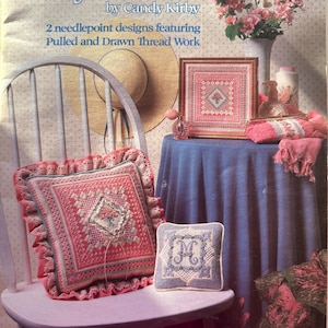 May include: A still-life image featuring needlepoint designs. The image includes a framed piece, pillows, and a leaflet titled "Fancy Threads" by Candy Kirby. The designs feature pulled and drawn thread work. A straw hat and flowers are also present.