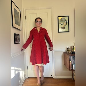 Puede incluir: Un abrigo rojo hasta la rodilla con un estampado floral. El abrigo tiene mangas largas y una botonadura frontal. La persona lleva un vestido color crema y tacones color beige. El abrigo es de estilo vintage.