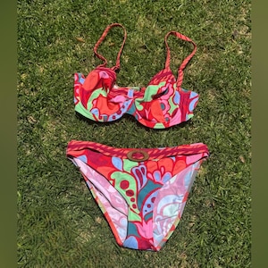 May include: A two-piece bikini set with a vibrant floral pattern in red, pink, green, and blue. The top has underwire support and adjustable straps. The bottoms feature a matching print and a decorative buckle.