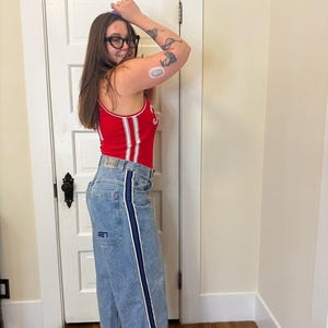 May include: A person wearing a red tank top with white stripes and light blue jeans with a dark blue stripe down the side. The person is wearing glasses and high heels.