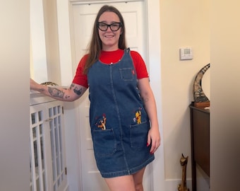 Könnte beinhalten: Jeans-Latzkleid mit gestickten Cartoonfiguren auf den Taschen. Das Kleid ist mittelblau mit einem roten Aufnäher auf der Brust. Ein rotes T-Shirt wird darunter getragen.