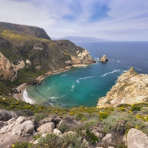 Puede incluir: Una vista a&eacute;rea de una cala aislada con una playa de arena y agua turquesa. La cala est&aacute; rodeada de acantilados rocosos y exuberante vegetaci&oacute;n verde. El cielo es azul brillante con nubes blancas.