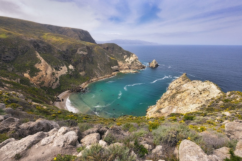 Puede incluir: Una vista a&eacute;rea de una cala aislada con una playa de arena y agua turquesa. La cala est&aacute; rodeada de acantilados rocosos y exuberante vegetaci&oacute;n verde. El cielo es azul brillante con nubes blancas.