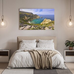 Puede incluir: Un interior de dormitorio con una cama blanca, una manta marr&oacute;n y una mesita de noche blanca. Una gran fotograf&iacute;a enmarcada de un paisaje costero cuelga sobre la cama.