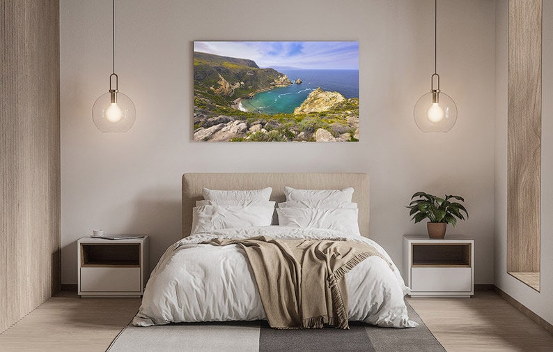 Puede incluir: Un interior de dormitorio con una cama blanca, una manta marr&oacute;n y una mesita de noche blanca. Una gran fotograf&iacute;a enmarcada de un paisaje costero cuelga sobre la cama.
