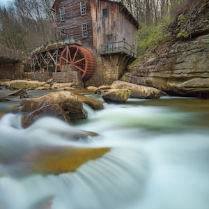May include: A rustic wooden watermill with a large red water wheel sits beside a rushing river. The mill is surrounded by rocks and trees, and the water is flowing quickly over the rocks.
