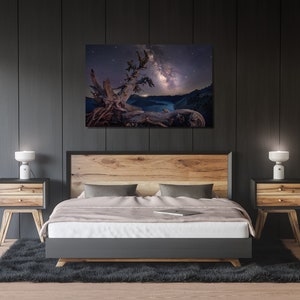 May include: A bedroom interior with a bed, two nightstands, and a framed photograph of a tree with the Milky Way galaxy in the background. The bed has a wooden headboard and a white duvet. The nightstands are made of wood and have two drawers each. The photograph is in a black frame and is hanging on a dark gray wall.