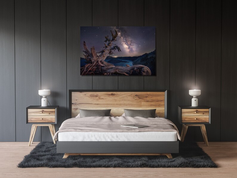 May include: A bedroom interior with a bed, two nightstands, and a framed photograph of a tree with the Milky Way galaxy in the background. The bed has a wooden headboard and a white duvet. The nightstands are made of wood and have two drawers each. The photograph is in a black frame and is hanging on a dark gray wall.