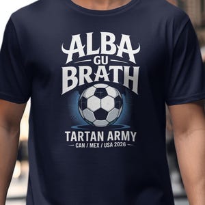 Puede incluir: Camiseta azul marino con un gráfico blanco y azul. El gráfico incluye el texto "ALBA GU BRATH" encima de un balón de fútbol y "TARTAN ARMY - CAN / MEX / USA 2026" debajo. La camiseta es de manga corta.