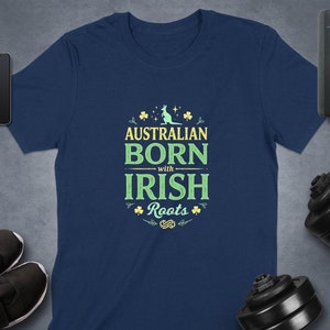 Puede incluir: Camiseta azul marino con el texto "AUSTRALIAN BORN with IRISH Roots" en verde y amarillo. El diseño incluye un canguro y tréboles. La camiseta se muestra con un smartphone, un smartwatch y equipo de entrenamiento.