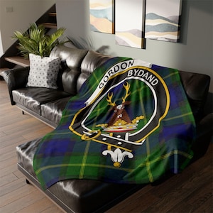 May include: A green, blue and yellow tartan throw blanket with a black crest featuring a stag and the text "Gordon Bydand".