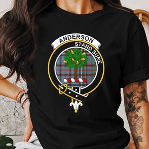 May include: Black t-shirt featuring a circular crest with the name "ANDERSON" and the motto "STAND SURE". The crest includes a tree, tartan pattern, and decorative elements. The shirt is a classic crew neck.