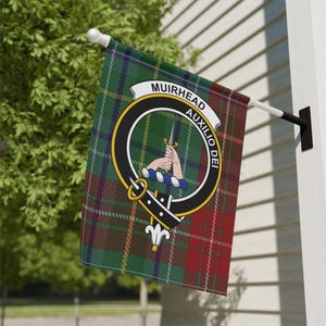 May include: A rectangular flag with a green, red, and black plaid pattern. The flag displays a circular emblem with the words "MUIRHEAD AUXILIO DEI". The emblem features a hand holding a dagger. The flag is mounted on a white pole.
