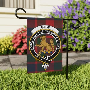 May include: A rectangular garden flag featuring a red, green, and dark blue tartan pattern. Centered is a circular crest with a gold cat, a black belt design, and the text "GOW" and "TOUCH NOT THE CAT BOT A GLOVE" in black lettering.