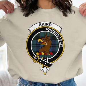 May include: Cream-colored sweatshirt featuring a circular emblem with the text "BAIRD DOMINUS FECIT". The emblem includes a brown eagle, a green and blue plaid background, and a black and gold border. A white banner with black text is above the emblem.
