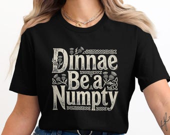 Puede incluir: Camiseta negra con la frase "Dinnae Be a Numpty" en una fuente desgastada de color blanquecino. El diseño incluye ilustraciones de cardos y nudos celtas. La camiseta es de cuello redondo.