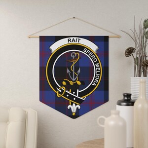 May include: A wall hanging featuring the Rait clan crest, with a blue, black, and red plaid background. The crest includes an anchor, a banner with the name "RAIT", and the motto "SPERO MELIORA". The banner is suspended from a wooden rod.