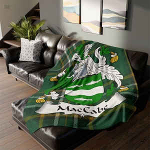 Könnte beinhalten: Grüne, gelbe und weiße karierte Plaid-Decke mit einem Wappen, das einen silbernen Helm, weiße Fische und grüne Blätter zeigt. Das Wappen ist von einem weißen Banner mit dem Text "MacCabe" umgeben.