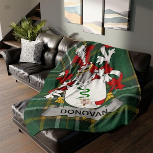 May include: A green and yellow plaid throw blanket with a family crest design. The crest features a helmet, red and white accents, and the name "DONOVAN" on a banner. The blanket is draped over a black leather sofa.