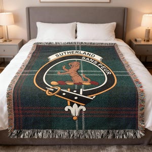 May include: A green and red plaid throw blanket with a fringed edge. The blanket features a crest with the words "SUTHERLAND SANS PEUR" and a brown lion. The blanket is displayed on a bed.