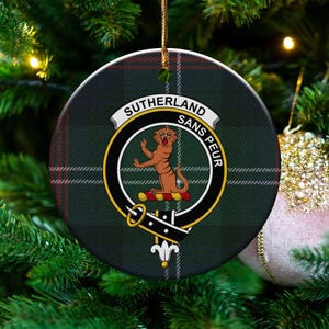 May include: Circular Christmas tree decoration with a green, black, and red tartan design. The ornament displays a crest with a tiger and the words "SUTHERLAND SANS PEUR". It is hanging on a Christmas tree.
