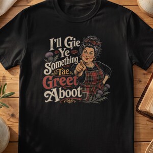 May include: A black t-shirt featuring a graphic illustration of a person wearing a plaid apron and pointing forward. The text reads, I'll Gie Ye Something Tae Greet Aboot, accompanied by images of purple thistle flowers on a dark background.