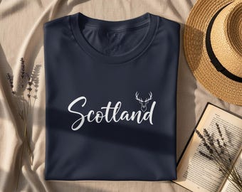 Puede incluir: Camiseta azul marino con la palabra "Scotland" en escritura blanca y un gráfico de cabeza de ciervo blanco. Un sombrero de paja y un libro abierto están junto a la camiseta sobre una superficie beige. También hay ramitas de lavanda secas.