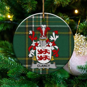 May include: A circular Christmas decoration with a green and yellow tartan design. It displays a family crest with a helmet, red and white details, and the name "CLANCY" on a banner. The ornament is suspended by a gold string.