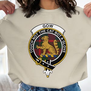 Op de afbeelding: Beige sweatshirt met een cirkelvormig embleem. Het embleem toont een tijgerillustratie en de tekst "GOW TOUCH NOT THE CAT BOT A GLOVE". Het embleem is omgeven door een zwarte rand en een tartan patroon.