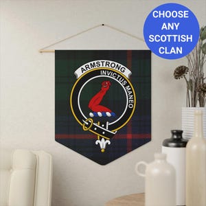 May include: A green, red, and black Scottish clan tapestry with the Armstrong clan crest. The crest features a red arm with a clenched fist and the Latin motto "INVICTUS MANEO". The tapestry is hanging on a wall with a wooden dowel rod.
